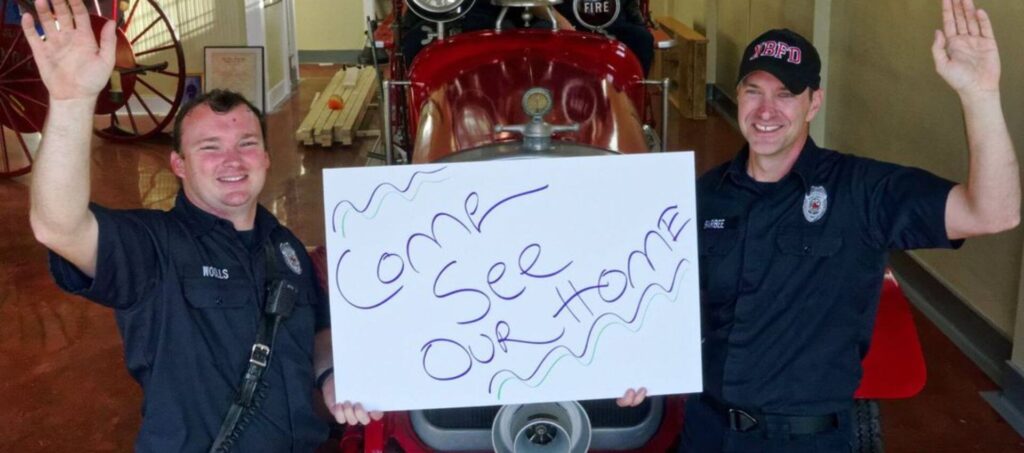 Two firemen inside the station house waving and holding a sign that says Come See Our Home