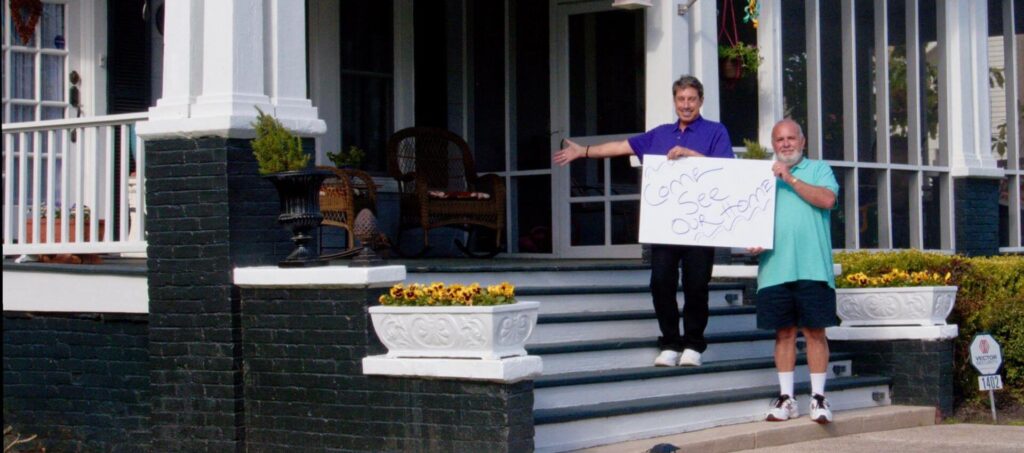 Randy Toran and Bob Lane standing on stairs holding sign Come See Our Home
