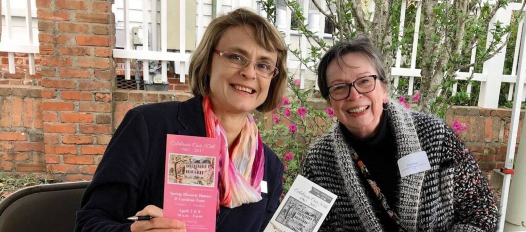 Two women smiling holding tickets on veranda surrounded by flowers